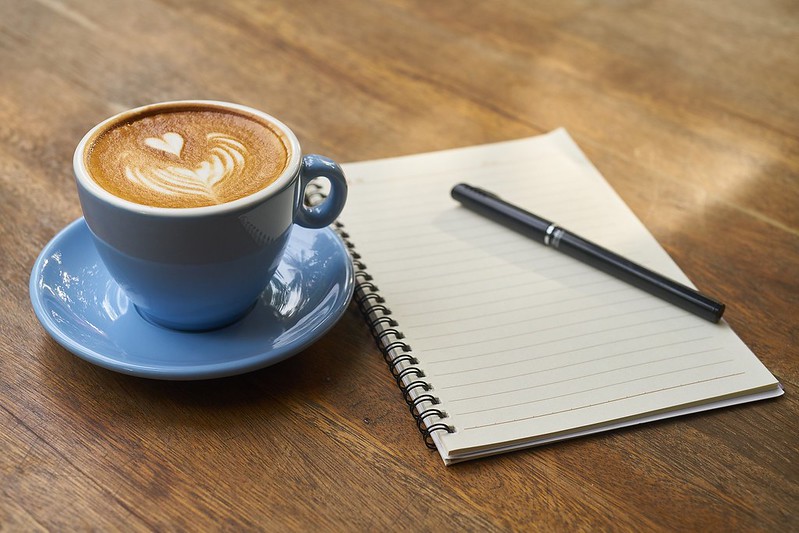 cappuccino in a blue demitasse next to a blank spiral-bound notebook with
closed pen resting on top